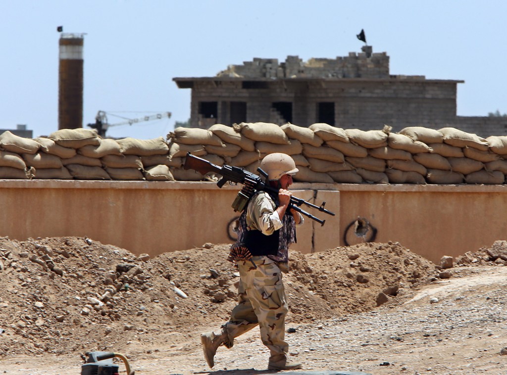 A Kurdish peshmerga fighter  walks to his base where two flags of the Islamic State are seen on a building, right, and water tower, left, at the front line with the al-Qaida-inspired militants in Tuz Khormato, 62 miles south of Kirkuk, northern Iraq, in June.