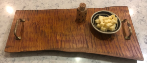 Serving tray made from live edge wood with metal handless. On the tray is a bowl of cheese and a toothpick holder.